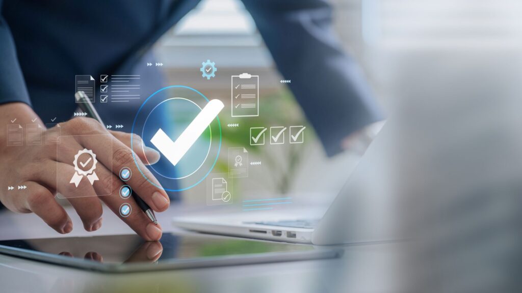 Hand mit Stift nutzt Tablet mit digitalen Abhaken-Symbolen und Qualitaetsmanagement-Icons im Buero