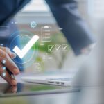 Hand mit Stift nutzt Tablet mit digitalen Abhaken-Symbolen und Qualitaetsmanagement-Icons im Buero