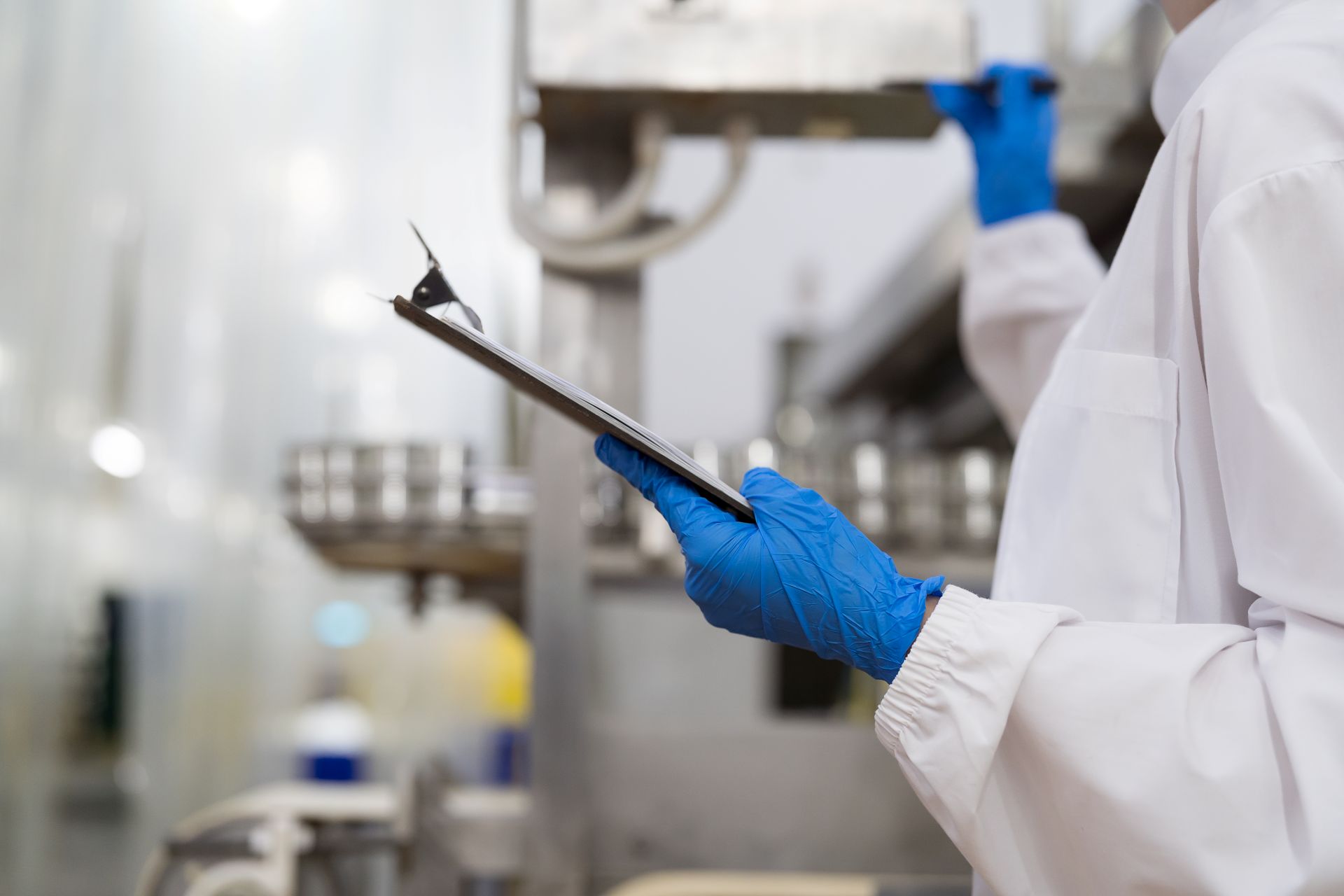 Person in weissem Schutzkittel und blauen Handschuhen fuehrt Qualitaetskontrolle mit Klemmbrett in einer modernen Produktionsanlage durch